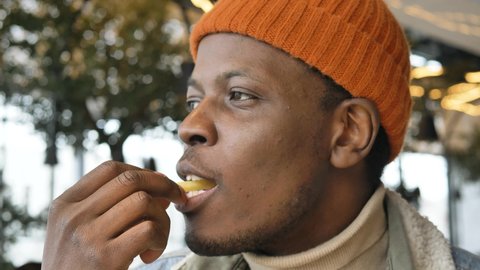 African hipster man in an orange hat eats delicious French fries and admires the taste in a fast food restaurant close-up. Harmful and unhealthy food.