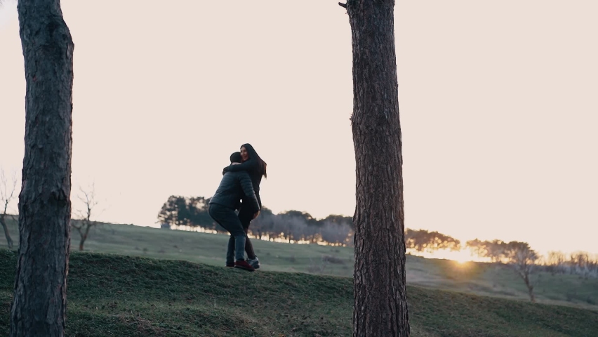 The young people in love, dressed in black, hug tightly, the man takes her in his arms and lifts her with much love, the woman smiles wide, happy, in the middle of nature in the light of the sunset