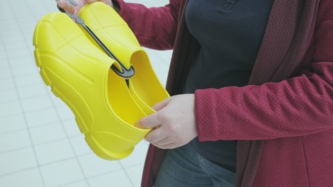 Pregnant woman in a store chooses yellow rubber galoshes for walking in rainy weather. Concept of buying new clothes, fashion, beauty and an active lifestyle while carrying child. Hands close up shot