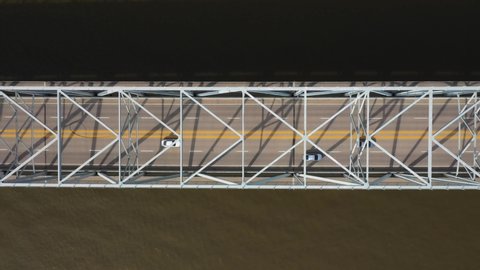 4K aerial overhead shot of a metal and concrete bridge as the camera floats left-to-right and lands on shore, still following the road.