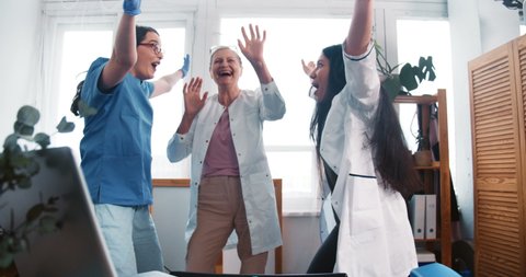 End of fighting COVID-19 pandemic. Three happy cheerful multiethnic female doctors dance celebrating big success at work
