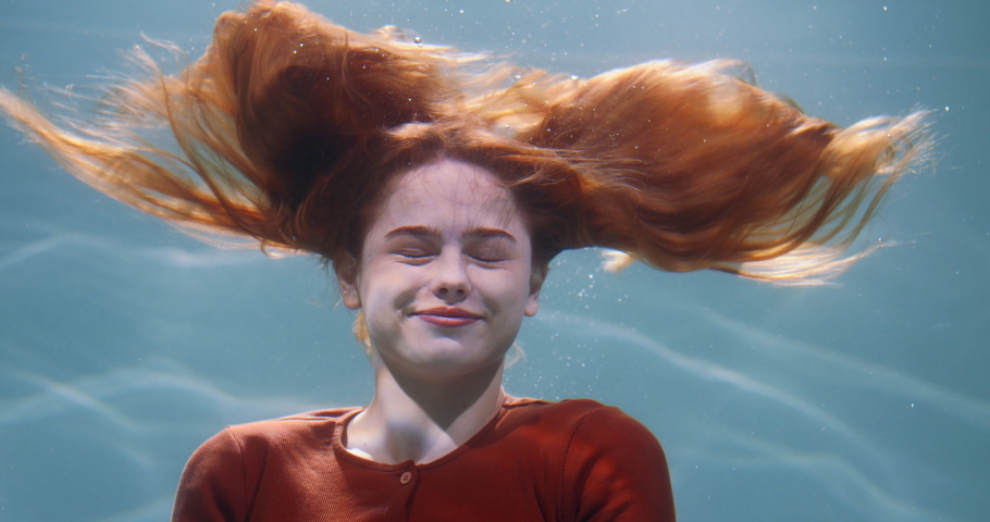 Amazing cinematic portrait of young beautiful redhead woman spreading hair, posing smiling deep under water slow motion.
