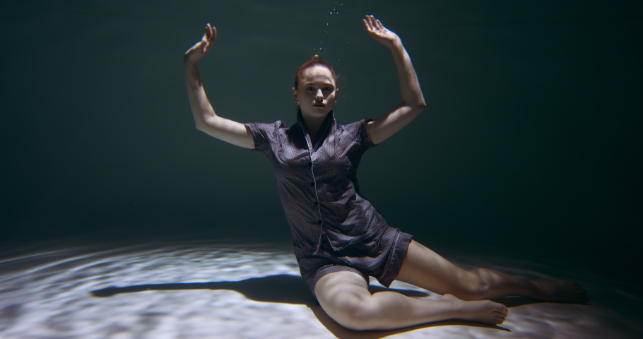 Young beautiful redhead woman wearing pyjamas falling down deep under water looking at camera in dark pool slow motion.