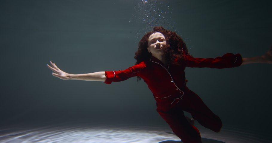 Amazing cinematic portrait of young beautiful woman in red pyjamas slowly sinking under water falling asleep slow motion