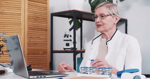 Senior healthcare professional woman in lab coat shows COVID-19 vaccine flask to remote patient via laptop webcam online