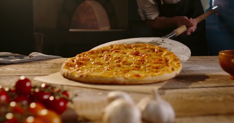 Senior chef taking hot pizza out of stone oven with peel and putting it on table. Experienced bakery worker cooking pizza with traditional recipe - small business 4k footage
