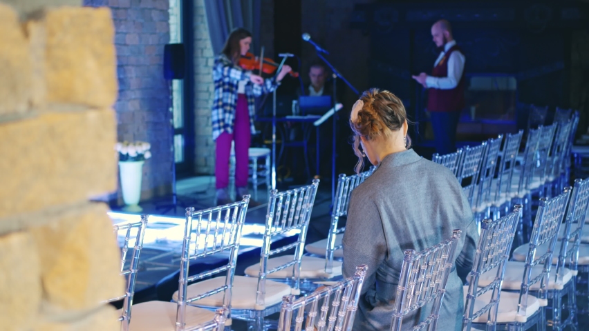 Preparing for the evening. Girl violinist rehearses the violin. There are chairs in front of the stage in the hall. The view from behind a woman in a gray dress. Rehearsing before the concert.