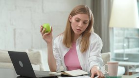 Focused woman sits at home on the desk and works using a laptop, eats an apple, manager on a remote work during self-isolation in quarantine, working with documents and reporting.