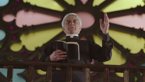 Low angle view of old Caucasian clergyman wearing black clothes and white collar standing in Christian church against stained-glass window with Bible in hands and preaching Sunday sermon