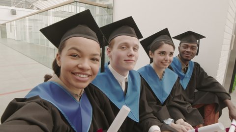 Medium shot of four diverse classmates wearing university graduate gowns and caps sitting together, looking at camera and waving their diplomas while making selfie