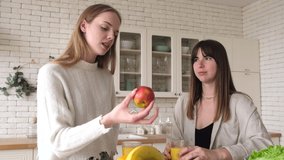 Two woman friends at home in kitchen having positive conversation talking about health and fresh fruits, diet, losing weight, hold apple  
