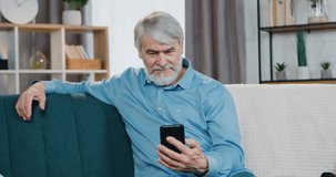 Handsome mature man relaxing on couch and using modern smartphone for video chat. Bearded male in casual clothes having online conversation while staying at home.