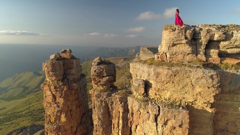 Drone orbit beautiful girl in long red dress stands dangerously on top of cliff rock. Incredible natural epic landscape Caucasus Elbrus region Bermamyt plateau travel landmark. Summer, clouds horizon