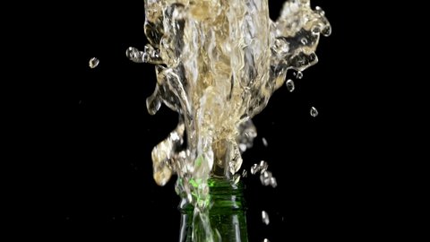 Macro shot of cap popping out of green glass bottle and explosion of splash carbonated beer. Amber liquid under pressure bursts out of bottle and fountains up. Black background. Close up. Slow motion.