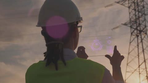 Woman power engineer in helmet working on futuristic tablet checking data of high-voltage electrical line at sunset. Ensuring uninterrupted supply and distribution of electricity to consumers.
