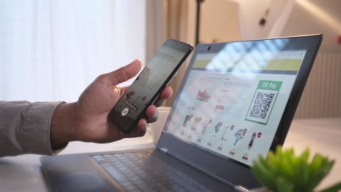 Handheld shot of an African-American male hand holding a mobile phone, using together with a laptop for online shopping, scanning QR code and paying with bitcoins via the mobile app