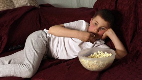 Lazy boy in casual white t-shirt lying on sofa and eating popcorn watching cartoons or movie on couch at home. Concept of cheerful children and family happiness