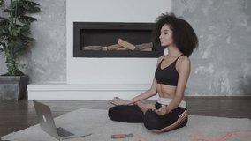 Beautiful young multiracial woman does yoga and strength training exercises on a mat in her living room. She follows an online exercise video on her laptop.