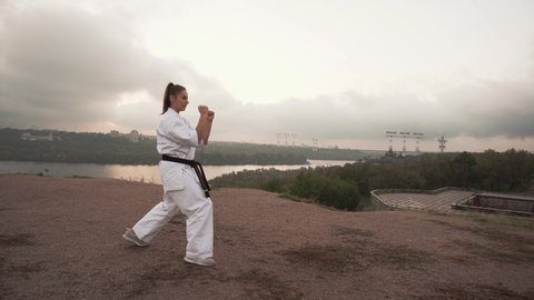 Translation: Kyokushinkai. karate girl in white kimono slowly makes 2 kicks in the air outdoors in the early morning