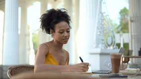 A female Afro-looking blogger writes down lessons in a diary. Sitting in a summer restaurant on a break. There is cocoa on the table