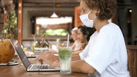 Remote work during a covid-19 pandemic. A young woman in a face mask working at a coffee place, takes off the face mask and sips matcha tea latte 