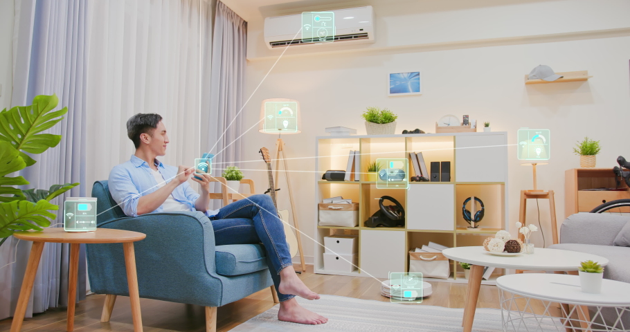 Asian man sitting on sofa and using smart home control app on mobile phone with augmented reality view of IOT connected objects in apartment