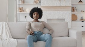 Bored lonely young African American woman sitting on couch in living room, holding TV remote control switch channels choosing programs, enjoying watching series on weekend at home, leisure pastime
