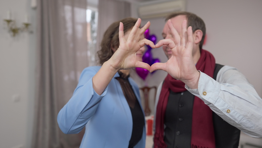 Couple hands forming heart shape with happy loving man and woman kissing and looking at camera at background. Cheerful Caucasian adult husband and wife celebrating Valentine