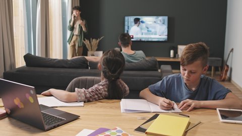 Waist-up shot of two elementary age kids having online lesson on laptop while their parents relaxing in background – mom talking on phone with friend and dad switching channels on tv