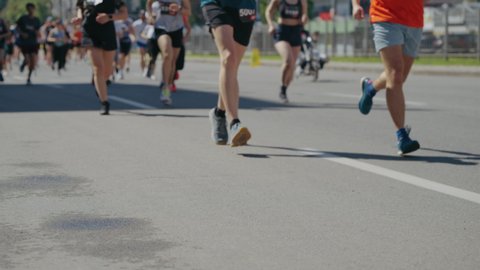 Legs of athletes running marathon on asphalt road in slow motion, focus on female legs, disabled person riding pumped wheelchair on background. Tracking shot partial view of sports event contestants