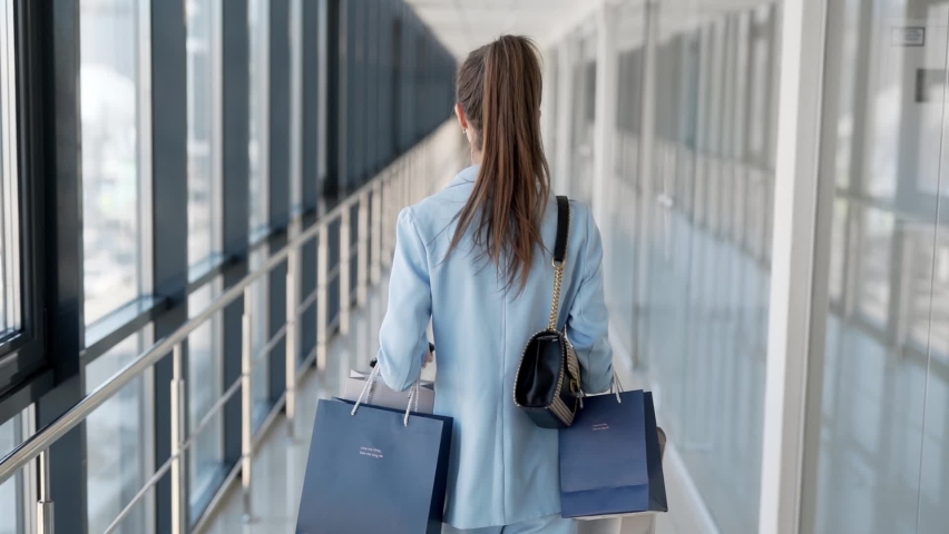 rear view Stylish young girl with packages walks down the glass corridor and smiles