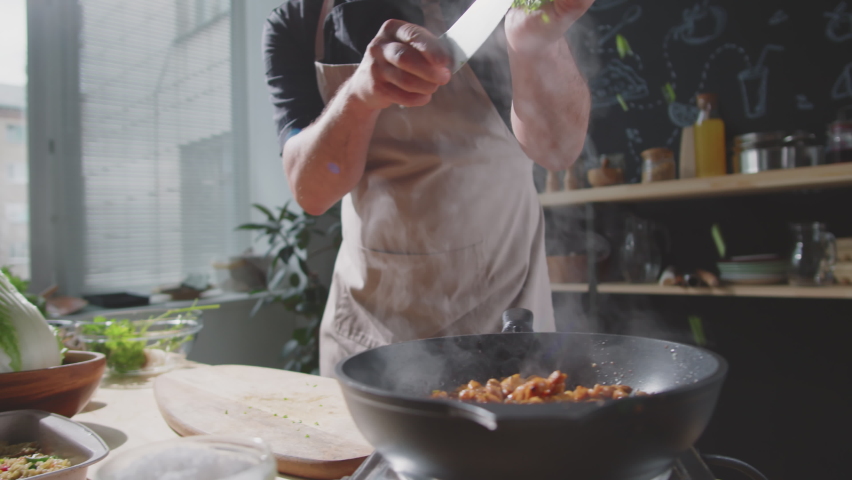 Midsection shot of male chef in apron adding chopped parsley to meat and tossing food on hot skillet while cooking meal in kitchen | Shutterstock HD Video #1072723847