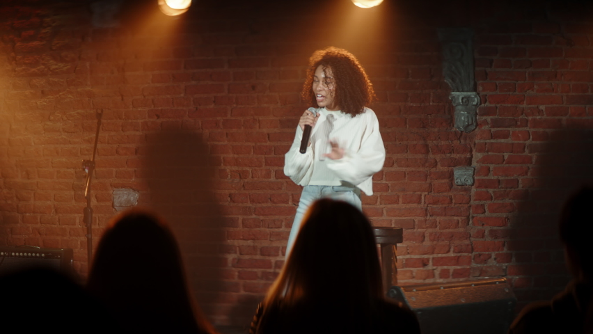 DOLLY SHOT Black African American young female comedian performing her stand-up monologue on a stage of a small venue. Shot with ARRI Alexa Mini LF with 2x anamorphic lens Royalty-Free Stock Footage #1072727297