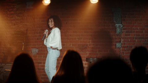 DOLLY SHOT Black African American young female comedian performing her stand-up monologue on a stage of a small venue. Shot with ARRI Alexa Mini LF with 2x anamorphic lens