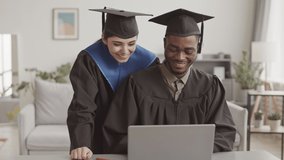 Medium shot of young mixed-race woman and African man both wearing graduation gowns and hats having online graduation ceremony at home using laptop