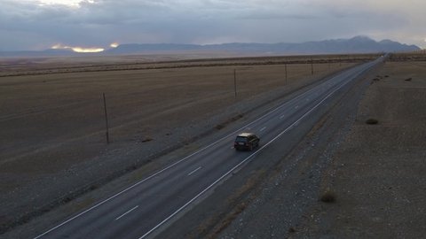 Automobile running on countryside roadway. Tracking shot drone view of car moving on asphalt road leading through hilly fields towards Altay mountains in cloudy evening