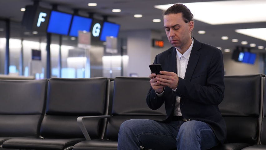 Man take look at smartphone, then put it in pocket of jacket. Portrait shot of passenger wasting time, waiting for boarding, sitting alone at terminal lounge Royalty-Free Stock Footage #1072737131