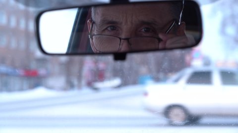An elderly good-natured man with glasses looks in the rear-view mirror, sitting in the car.