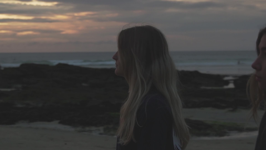 Two silhouetted young girls walk in slow motion down a beach past a golden sunset with a rocky coastline in the background.