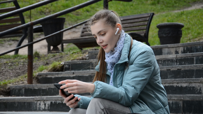 Side view of Cheerful young girl student sitting on the steps listens to music and smiles with