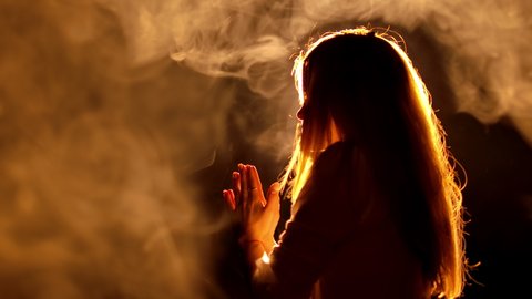 A blonde Christian girl staying in church and praying. Light source reflection camera lens making amazing mystic view. Incense smoke around the woman isolated on black background. 