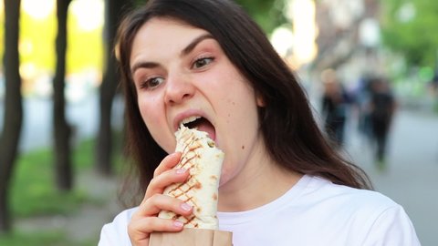 Brunette woman overeats shawarma on a city street. Street fast food pita roll with meat and vegetables.