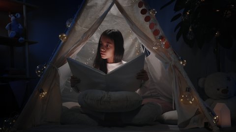 Smart girl with long black hair in cute decorative tent looking to book with flashlight. Pretty teenager sitting on floor while reading . Concept of leisure and free time