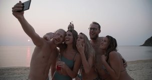 Young caucasian men and women use smartphone to video call on the beach. Group of friends in swimwear enjoy talking and drinking party at sunset in summertime. Travel, vacation and friendship concept.