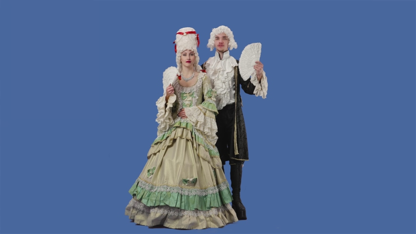 Portrait of courtier lady and gentleman in historical vintage costumes and wig is waving a fan. Young woman and man posing in studio with blue screen background. Full lenght. Slow motion. Royalty-Free Stock Footage #1072758971