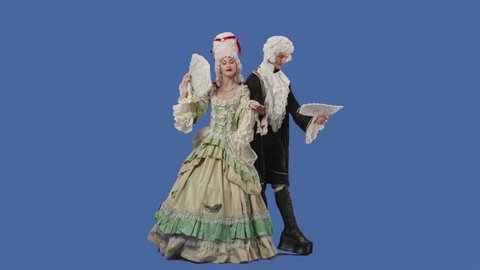Portrait of courtier lady and gentleman in historical vintage costumes and wig is waving a fan. Young woman and man posing in studio with blue screen background. Full lenght. Slow motion.