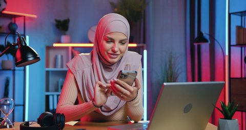 Beautiful smiling happy modern young muslim woman in hijab uses her smartphone while making break in her work on laptop in home office in the evening