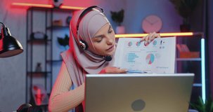 Adorable positive confident skilled muslim young woman in hijab explaining charts via video meeting on laptop sitting in home office in the evening