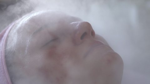 Close up portrait of young woman with eyes closed having thermal steam treatment on face. Girl wearing white gown laying on couch in spa