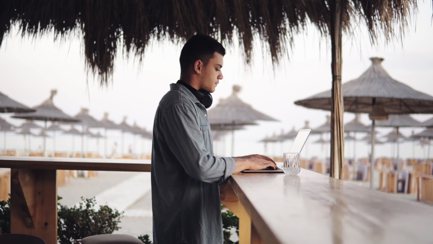 Young successful developer man working with laptop in the beach bar by the sea. Freelance work in tropical country Royalty-Free Stock Footage #1072761353